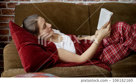 Portrait of young woman in pajamas lying on couch and reading book. People resting at home, hobby, education, female leisure Portrait of young woman in pajamas lying on couch and reading book. People resting at home, hobby, education, female leisure 106182160