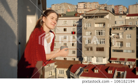 Beautiful woman in plaid drinking coffee while looking through window on city. People relaxing, resting at home, beautiful cityscape. 106182194