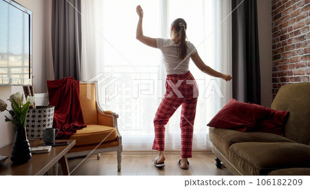 Funny shot of happy woman wearing pajamas and dancing against big window in living room. People relaxing at home, having fun, positive emotions and fun. Funny shot of happy woman wearing pajamas and dancing against big window in living room. People relaxing at home, having fun, positive emotions and fun. 106182209