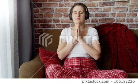 Young woman wearing headphones meditating on sofa in living room. Concept of relaxation, healthy lifestyle, resting at home and practicing yoga. 106182220