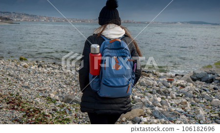 Young female hiker with backpack and thermos walking on the rocky sea beach at cold cloudy and windy day. Concept of hiking adventure, exploration, tourism and journey. 106182696