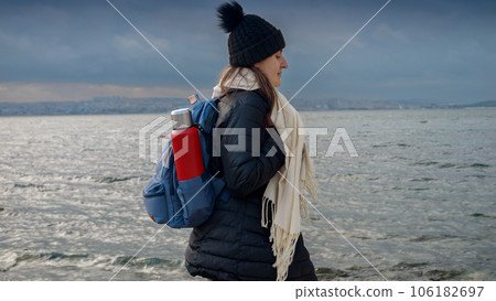Female hiker, equipped with a backpack and thermos, exploring a rocky sea beach on a cold, cloudy day. Addition to any travel, tourism or adventure-themed project. 106182697