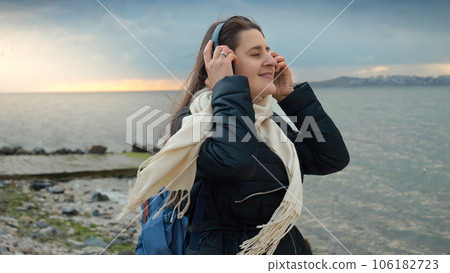 A smiling woman carrying a backpack walks along the beach while listening to music on a windy, chilly day. Essence of adventure, travel, and enjoyment. The perfect choice for travel-themed videos or A smiling woman carrying a backpack walks along the beach while listening to music on a windy, chilly day. Essence of adventure, travel, and enjoyment. The perfect choice for travel-themed videos or 106182723
