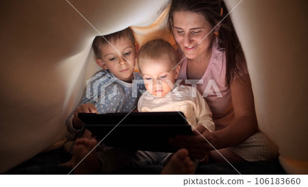 Happy family in pajamas browsing internet and watching video on tablet computer in bed under blanket. Family having time together, parenting, happy childhood and entertainment. Happy family in pajamas browsing internet and watching video on tablet computer in bed under blanket. Family having time together, parenting, happy childhood and entertainment. 106183660
