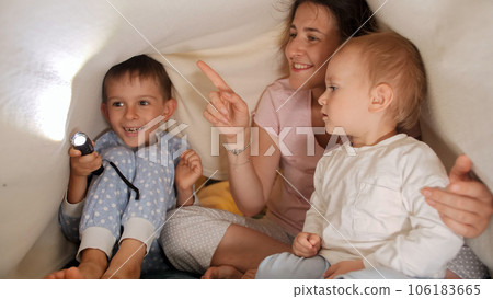 Cute baby boy with brother and mother playing with flashlight and shadows under blanket in bed. Family having time together, parenting, happy childhood and entertainment. Cute baby boy with brother and mother playing with flashlight and shadows under blanket in bed. Family having time together, parenting, happy childhood and entertainment. 106183665