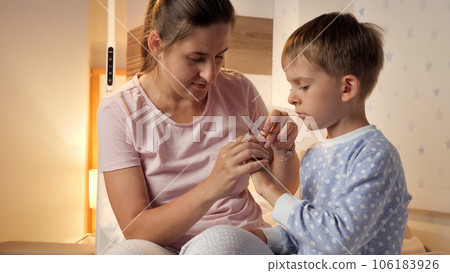 Young caring mother cutting fingernails to her son wearing pajamas in bedroom Parenting, child hygiene and healthcare, going to sleep. 106183926