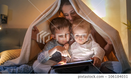 Smiling family hiding under blanket at night and using tablet computer. Family having time together, parenting, happy childhood and entertainment. 106184031