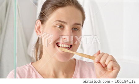 Smiling brunette woman brushing teeth with wooden toothbrush in the bathroom. Concept of teeth health, self checking mouth and oral hygiene. 106184112