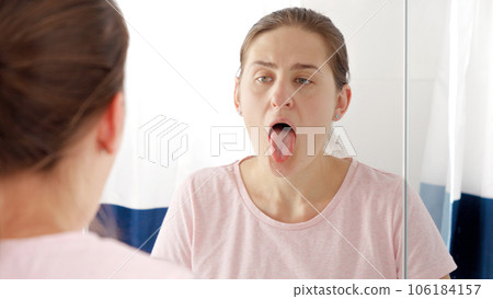 Young woman standing at mirror and checking her tongue for plaque and diseases. Concept of teeth health, self checking mouth and oral hygiene. 106184157