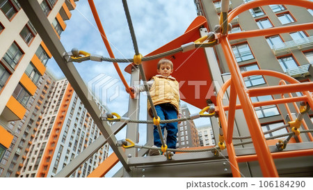 View from the ground on little boy walking over the rope bridge at playground. Active child, sports and development, kids playing outdoors View from the ground on little boy walking over the rope bridge at playground. Active child, sports and development, kids playing outdoors 106184290