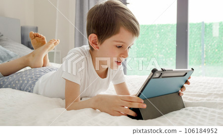 Little boy lies in bed with a tablet computer enjoying his screen time. Child using gadget, education and development. Little boy lies in bed with a tablet computer enjoying his screen time. Child using gadget, education and development. 106184952