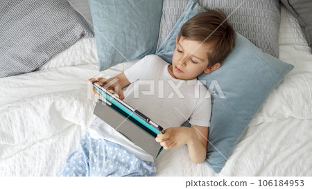 Little boy is lying on his bed, using a tablet computer during the day. Child using gadget, education and development. Little boy is lying on his bed, using a tablet computer during the day. Child using gadget, education and development. 106184953