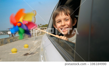 Smiling boy looking through open car window and holding colorful spinning pinwheel because of blowing wind. Concept of children travel, journey, tourism, holiday, weekend and vacation. Smiling boy looking through open car window and holding colorful spinning pinwheel because of blowing wind. Concept of children travel, journey, tourism, holiday, weekend and vacation. 106184990