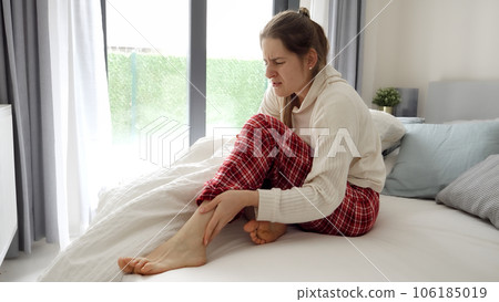 Young woman feeling cramp in leg lying in bed and massaging feet. Concept of injury, health problems, pain and trauma. Young woman feeling cramp in leg lying in bed and massaging feet. Concept of injury, health problems, pain and trauma. 106185019