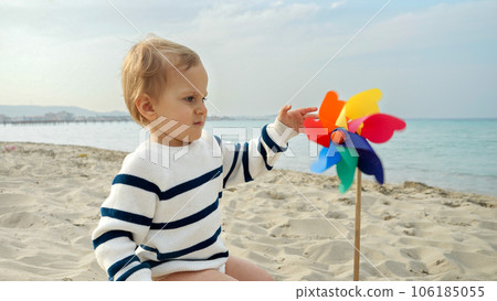 Portrait of cute baby boy sitting on the sandy sea beach and playing with colorful pinwheel. Concept of child happiness during summer vacation or weekend. Portrait of cute baby boy sitting on the sandy sea beach and playing with colorful pinwheel. Concept of child happiness during summer vacation or weekend. 106185055