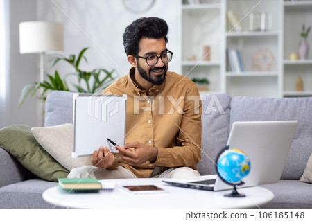 Young male teacher teaching remotely from home, hispanic using laptop for video call and communication and teaching students remotely, explaining new school material smiling. 106185148