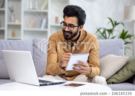 Young hispanic male student studying at home sitting on sofa, watching online course using laptop for video call, writing data in notebook, happy in living room. Young hispanic male student studying at home sitting on sofa, watching online course using laptop for video call, writing data in notebook, happy in living room. 106185150