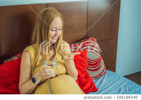 Prenatal Vitamins. Portrait Of Beautiful Smiling Pregnant Woman Holding Pill Box and a glass of water, Taking Supplements For Healthy Pregnancy While Sitting On Couch At Home, Free Space 106186540