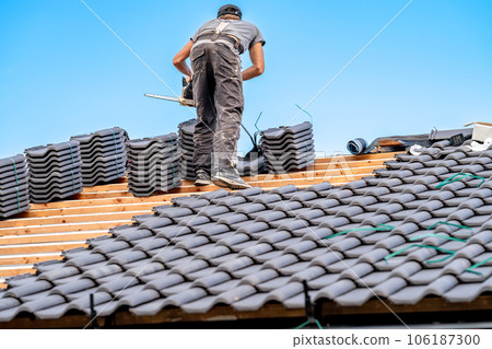 laborer with a chainsaw on the roof of a family house, building a roof from fired ceramic tiles 106187300