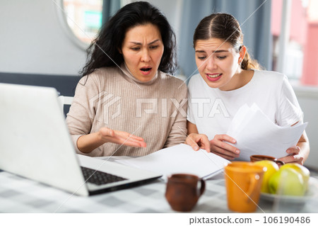 Two women with financial documents near laptop sitting at table at home 106190486