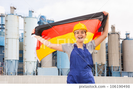 Happy girl in work clothes and hardhat with flag of germany standing in front of industrial scenery 106190498