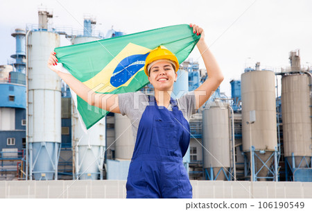 Happy girl in work clothes and hardhat with flag of brazil standing in front of industrial scenery Happy girl in work clothes and hardhat with flag of brazil standing in front of industrial scenery 106190549