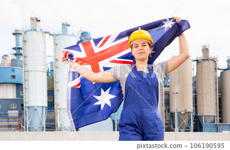Young female engineer in helmet waving state flag of Australia while standing in front of big tanks at chemical plant 106190595