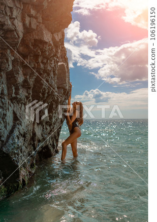 Woman travel sea. Attractive blonde woman in a black swimsuit enjoying the sea air on the seashore around the rocks. Travel and vacation concept. 106196150
