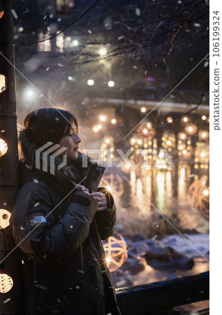 A woman standing in front of a bamboo lantern with the hot spring light in the background while the light snow falls 106199324