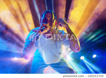 Rap musician. Young artistic man with dreads performing on stage over multicolored neon lights. Concert at night club 106201330