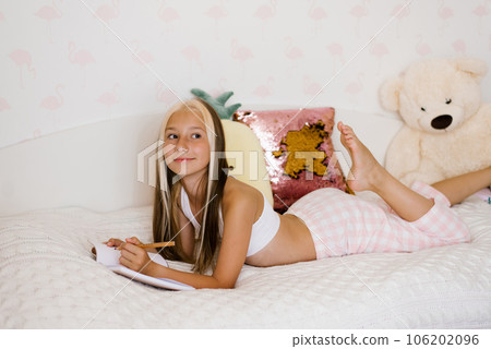 Cute Caucasian teen girl reads her diary sitting on the bed in sleeping clothes and makes notes in it Cute Caucasian teen girl reads her diary sitting on the bed in sleeping clothes and makes notes in it 106202096