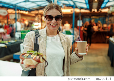 Smiling woman tourist is having a takeaway fast food lunch on food market and drink coffee Smiling woman tourist is having a takeaway fast food lunch on food market and drink coffee 106202191