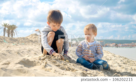 Two boys sitting on the sandy sea beach and playing with toy cars. Concept of tourism, travel, summer vacation Two boys sitting on the sandy sea beach and playing with toy cars. Concept of tourism, travel, summer vacation 106202215