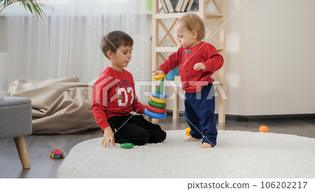 Little baby boy assembling colorful toy pyramid with his older brother in living room. Concept of child education, baby development, family playing games. Little baby boy assembling colorful toy pyramid with his older brother in living room. Concept of child education, baby development, family playing games. 106202217