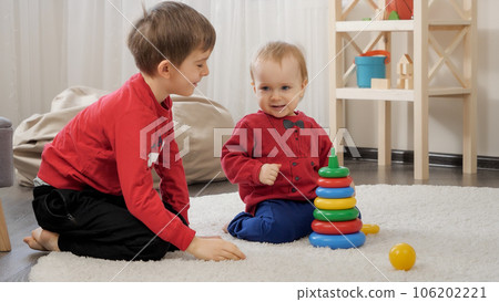 Smiling older boy playing with his little baby brother in toys on carpet at living room Smiling older boy playing with his little baby brother in toys on carpet at living room 106202221