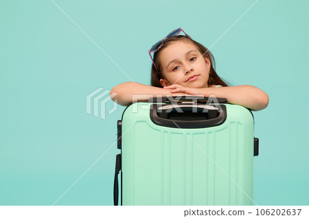 Lovely little girl traveler going for weekend getaway, looking at camera, posing with blue suitcase on studio background 106202637
