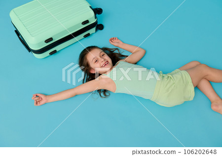 Mischievous little child girl with suitcase, lying on blue background, expressing positive emotions, looking at camera Mischievous little child girl with suitcase, lying on blue background, expressing positive emotions, looking at camera 106202648