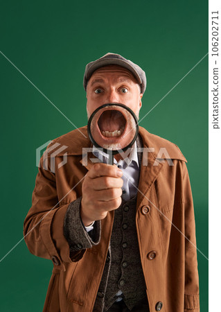 Half-length portrait of funny man, detective wearing vintage hat, suit with magnifying glass near face throught which can see his open mouth. 106202711