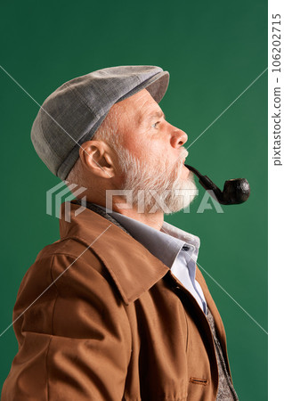 Portrait of side view confident and responsible man, detective wearing vintage hat, elegance suit with pipe in mouth on green color background. Portrait of side view confident and responsible man, detective wearing vintage hat, elegance suit with pipe in mouth on green color background. 106202715