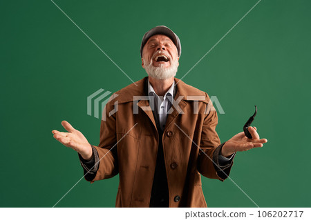 Portrait of positive, contented and happy man, detective wearing vintage hat, elegance suit with pipe in mouth on green color background. Portrait of positive, contented and happy man, detective wearing vintage hat, elegance suit with pipe in mouth on green color background. 106202717