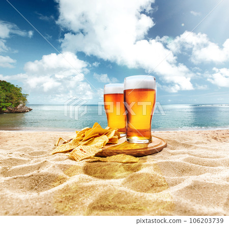 Two glasses of foamy, chill, lager beer with chips appetizers on sand against ocean and blue sky background 106203739