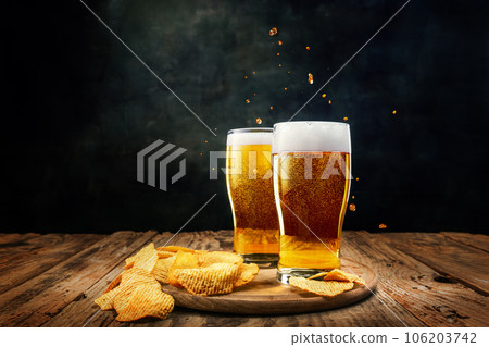 Two glasses of foamy, chill, lager beer with chips appetizers on wooden table against dark background Two glasses of foamy, chill, lager beer with chips appetizers on wooden table against dark background 106203742