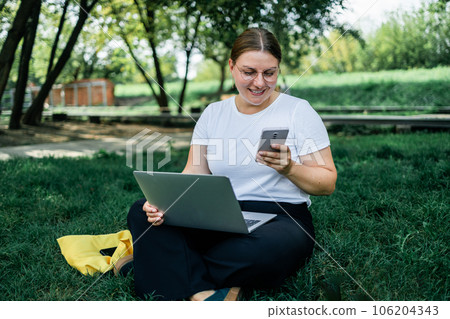 Happy Caucasian student freelancer woman rest use laptop pc computer talk by mobile cell phone look, sitting on grass outdoors in spring park. She is keyboarding on laptop. Freelance work concept. Happy Caucasian student freelancer woman rest use laptop pc computer talk by mobile cell phone look, sitting on grass outdoors in spring park. She is keyboarding on laptop. Freelance work concept. 106204343