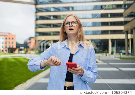 Angry irritated young business woman holding mobile phone and looks at his watch at the blurry business center building 106204374