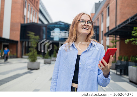Cheerful happy stylish businesswoman using smartphone online app while standing near the mall. Happy woman in a blue shirt walks down the street looking at her smartphone.An exciting application used 106204375