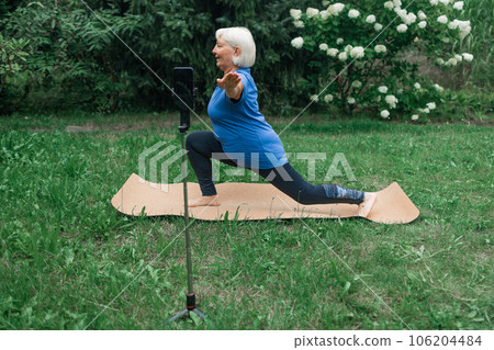 Fit mature 50s female sitting on yoga mat and talking at smartphone camera, making video content for followers, looking at webcam on mobile phone on tripod outdoors. Sports Blogger concept.  106204484