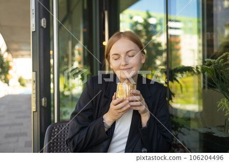 Close up of young female in business casual wear drinking cup of coffee while sitting at small table and enjoying time in street cafe. 106204496