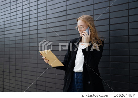 Portrait of corporate woman answers phone call while walking to the office building. Businesswoman calling someone, holding documents. 106204497