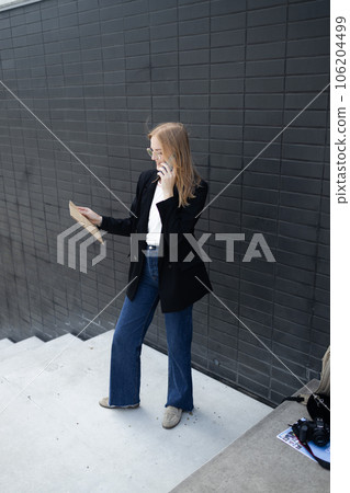 Portrait of corporate woman answers phone call while walking to the office building. Businesswoman calling someone, holding documents. 106204499