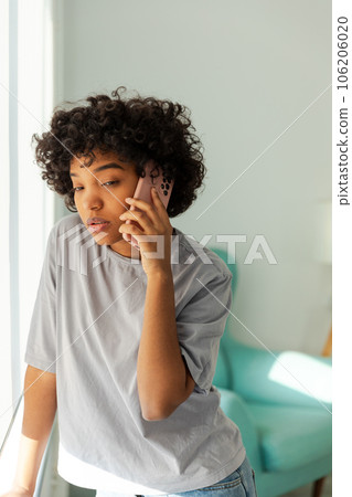 African american girl talking on smartphone at home indoor. Young woman with cell phone chatting with friends. Smiling teen girl making answering call by cellphone. Lady having conversation by mobile African american girl talking on smartphone at home indoor. Young woman with cell phone chatting with friends. Smiling teen girl making answering call by cellphone. Lady having conversation by mobile 106206020